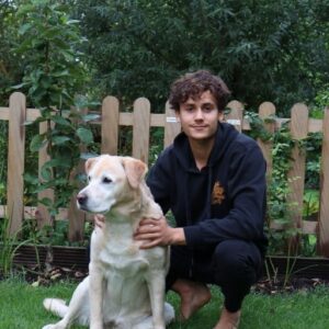 A photo of a young man with brown hair and brown eyes squatting next to a golden retriever in a green garden, the man is in a black tracksuit and looking into the camera, there is a wooden fence behind the man and the dog