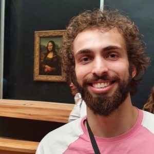 Photo of a young man with brown hair and blue eyes in a pink t-shirt smiling in front of the Mona Lisa painting